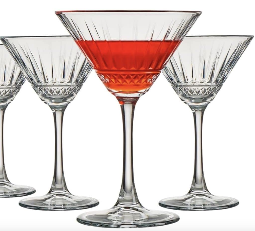 Four crystal martini glasses, one filled with a vibrant red liquid, are elegantly displayed. The centered glass hints at easy summer mocktail recipes, showcasing its striking hue. Each glass features intricate etched designs, adding a touch of sophistication to the scene.